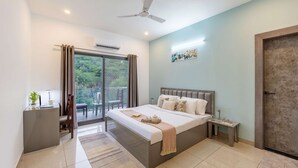 A modern bedroom with a large bed, a desk, a chair, a small table, and a balcony with a view of greenery.