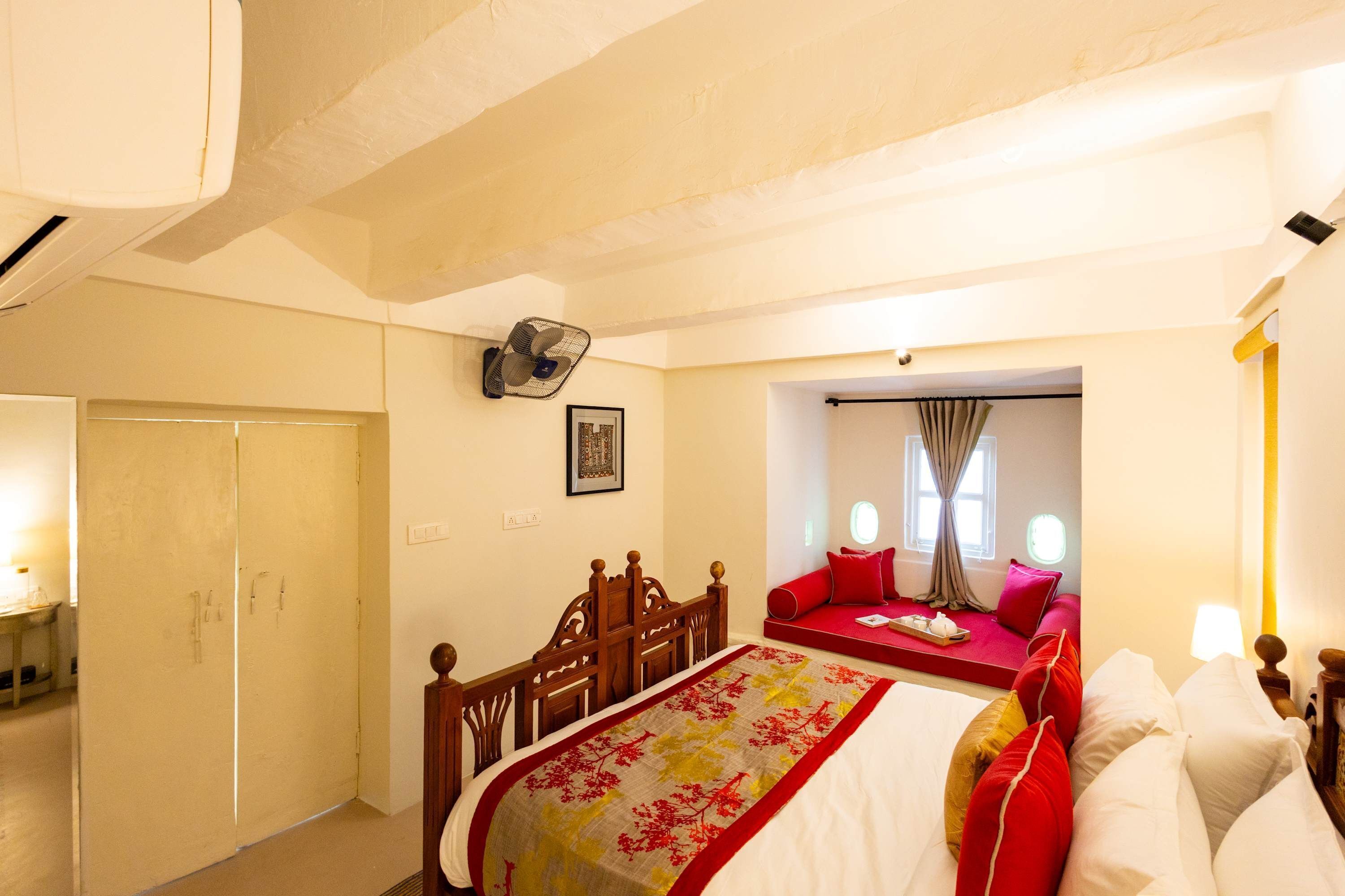 A bedroom with a bed, a wooden chair, a bedside table, a wall-mounted fan, and a window with curtains.