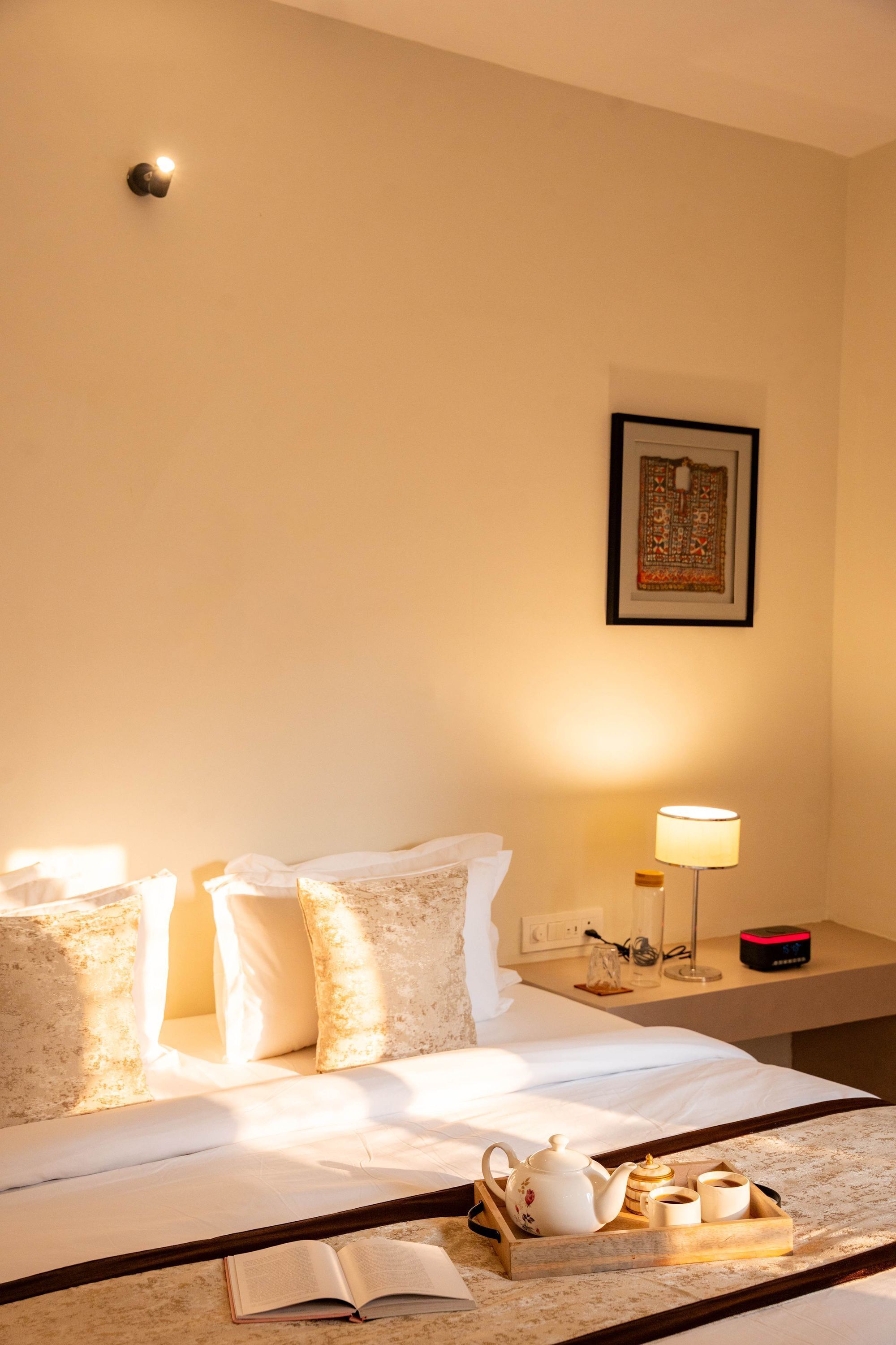 Une chambre d’hôtel avec un lit, des oreillers, une table de chevet avec une lampe, un tableau encadré et un plateau avec une théière et des tasses.