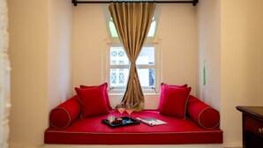 Une pièce chaleureuse avec un canapé rouge, deux coussins rouges, un plateau avec des verres et une bougie, et une fenêtre avec des rideaux.