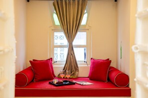 A cozy room with a red sofa, two red cushions, a window with a curtain, and a tray with glasses and a magazine.