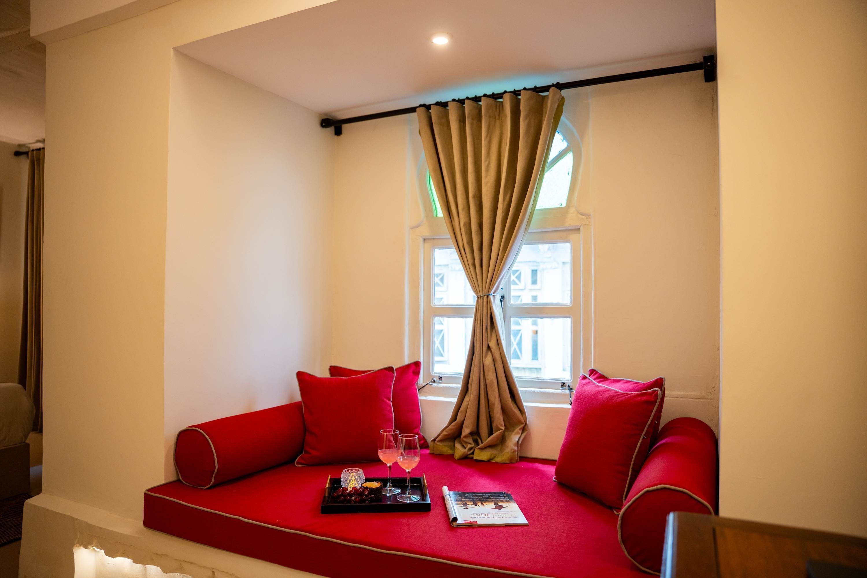 A cozy seating area with a red sofa, a window with beige curtains, and a tray with a drink and a book.