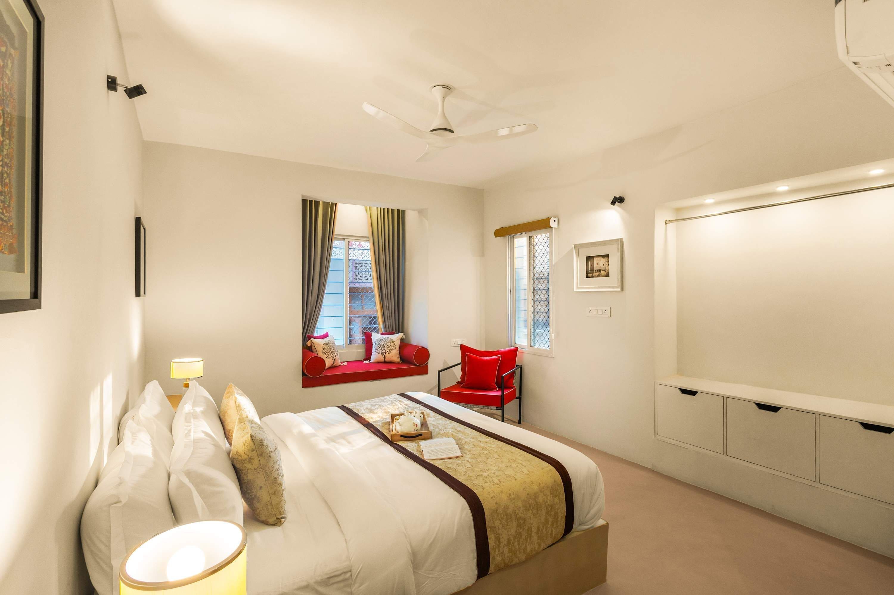 A hotel room with a bed, a window with curtains, a red chair, a bedside table with a lamp, and a wardrobe.