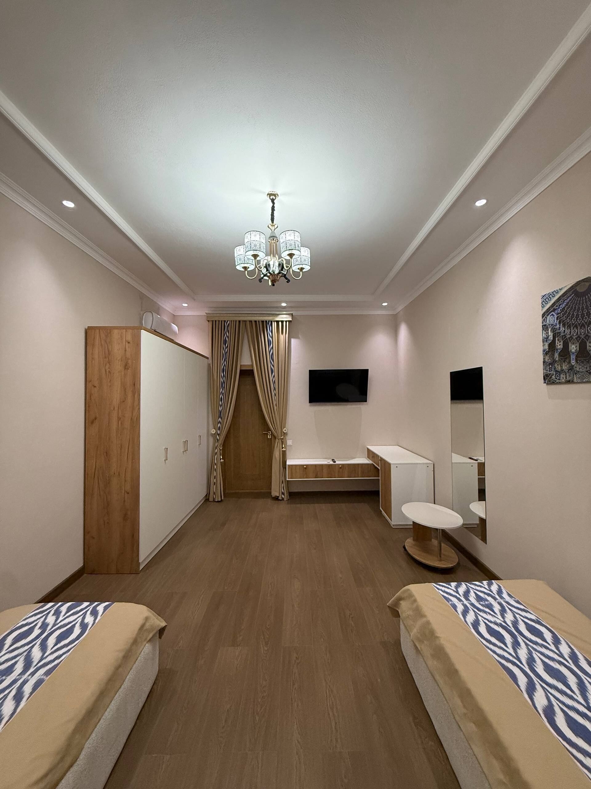 A hotel room with a wooden wardrobe, a vanity with a mirror, a bed with a patterned bedspread, and a chandelier.