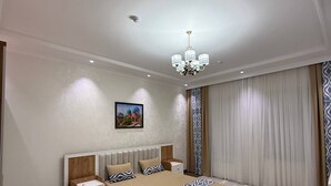 A hotel room with a large bed, a nightstand, a bedside table, a chandelier, a painting on the wall, and a patterned rug.