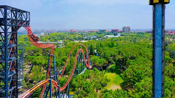 A roller coaster with multiple tracks, surrounded by greenery and urban buildings in the background.
