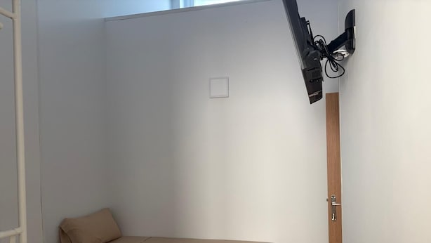 Pieni huone, jossa on yksi sänky, puinen pääty, musta tuoli, pieni pöytä, jossa on kaukosäädin, ja seinälle asennettu televisio.