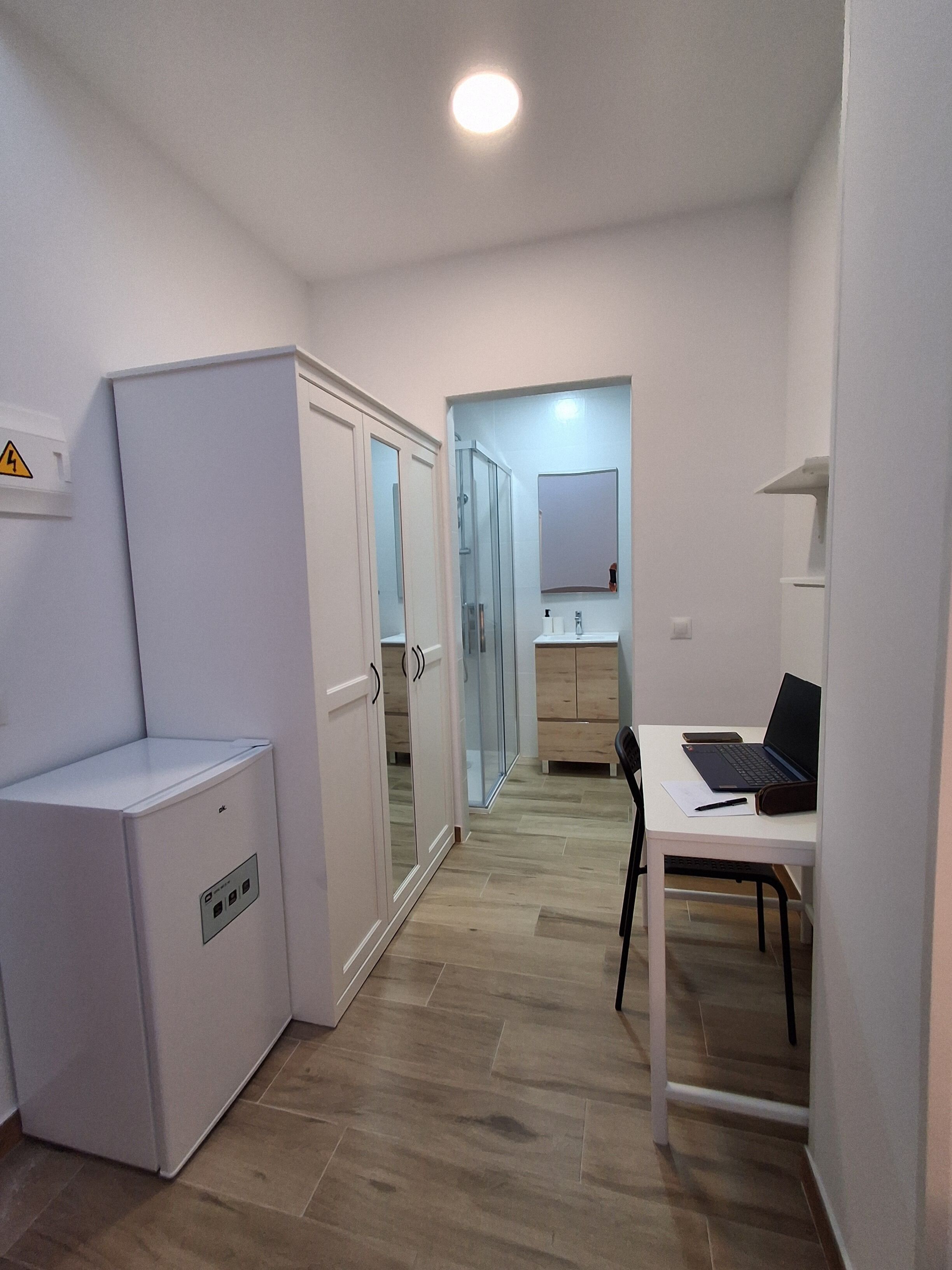 Een kleine kamer met een witte garderobe, een klein tafeltje met een laptop erop en een spiegel aan de muur.