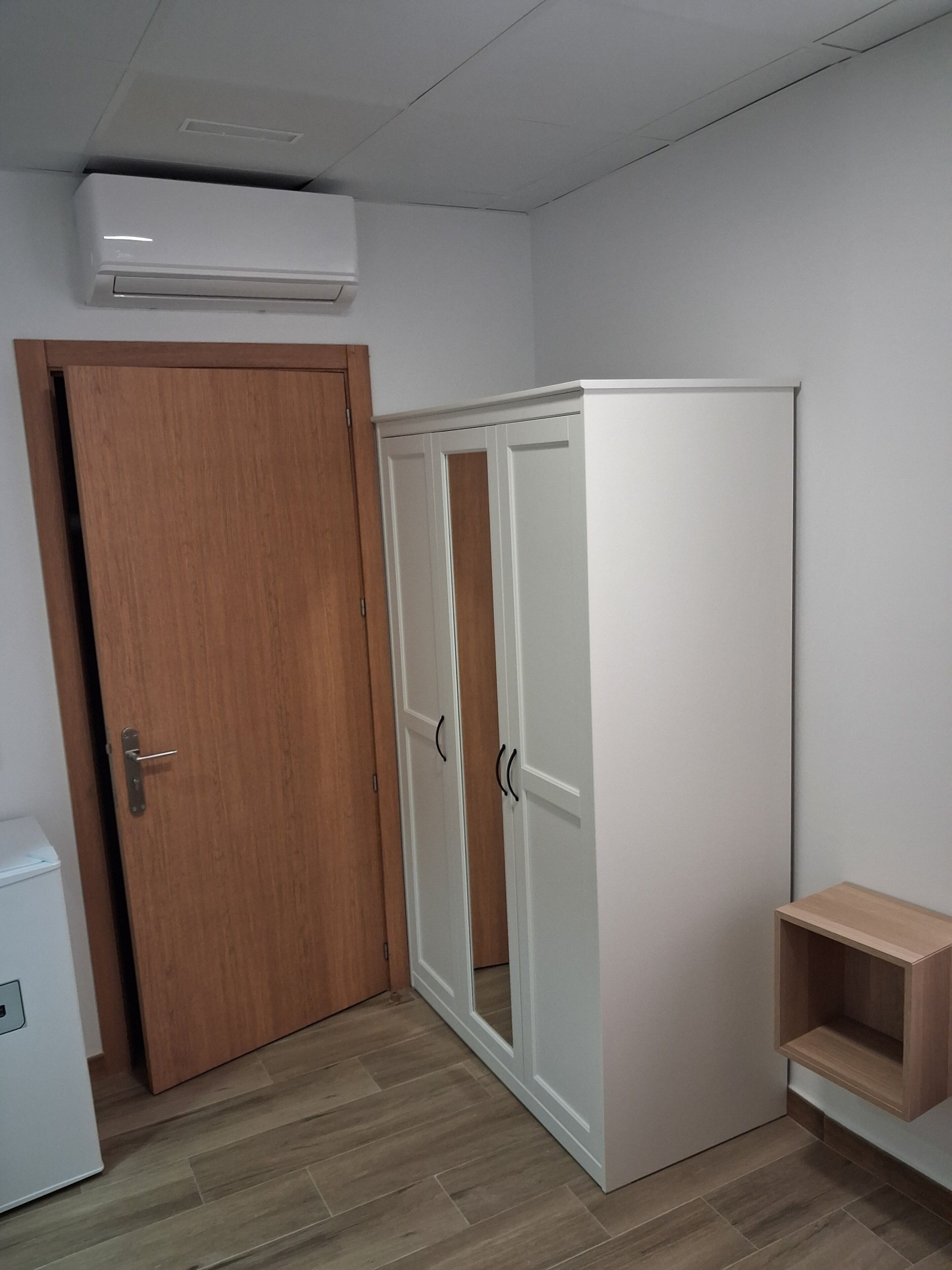 A room with a wooden door, a white wardrobe with two doors, and a small wooden shelf.