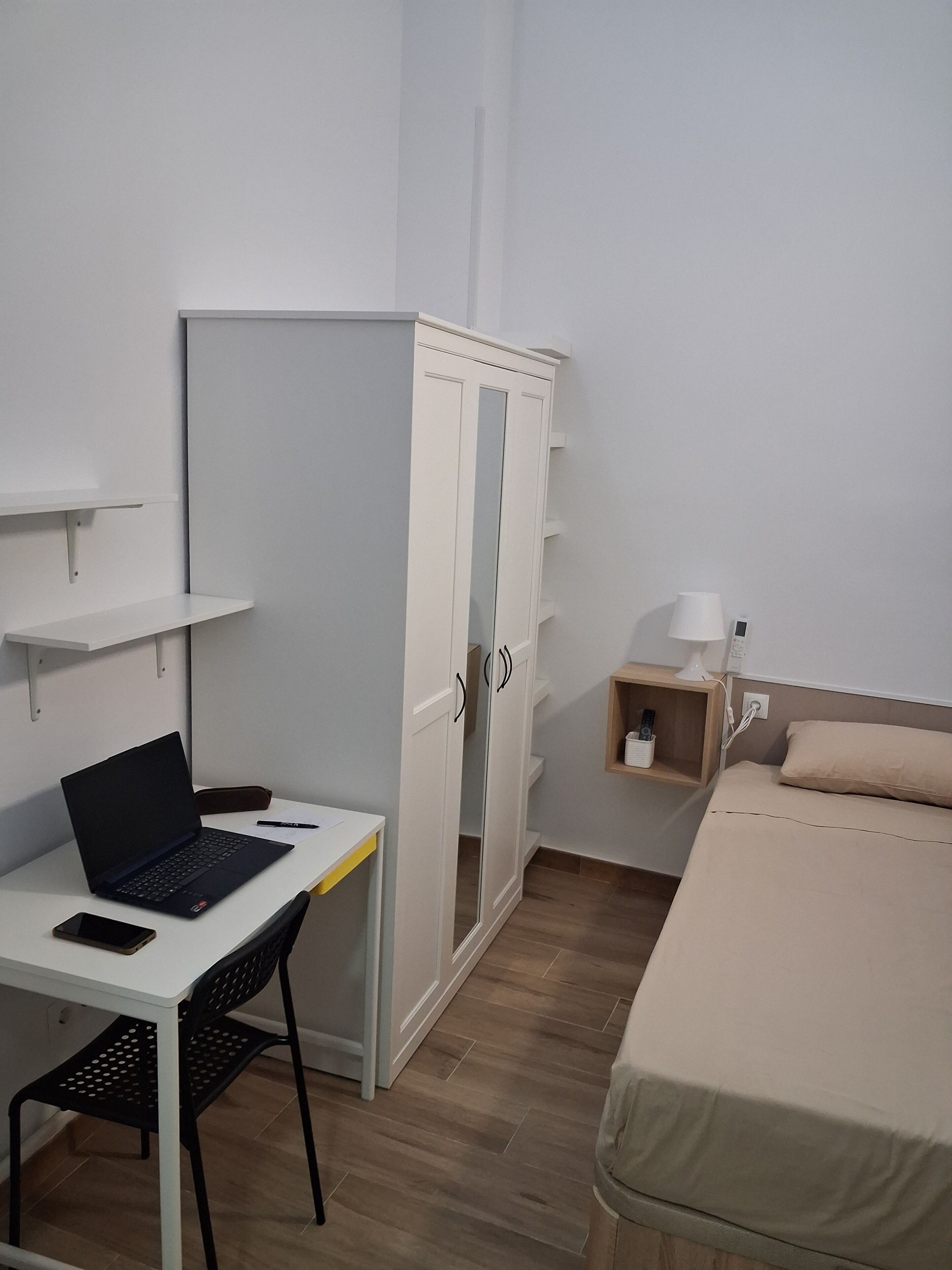 A bedroom with a bed, a desk with a laptop, a wardrobe, and a bedside table with a lamp.