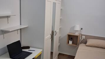 A bedroom with a bed, a desk with a laptop, a wardrobe, and a bedside table with a lamp.