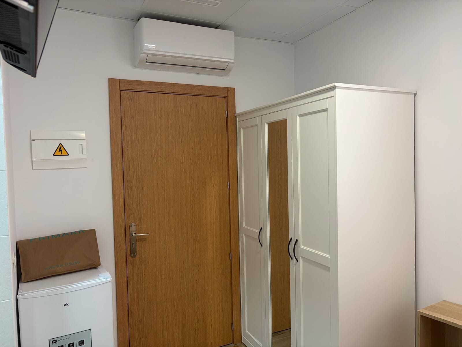 Een kamer met een houten deur, een witte garderobe, een witte airconditioning en een klein wit kastje met een bruine tas erop.