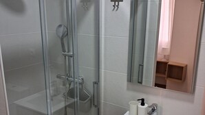 A modern bathroom with a glass-enclosed shower, a white sink with a mirror above it, and wooden cabinetry. - House Amara y Pablo (Madrid)