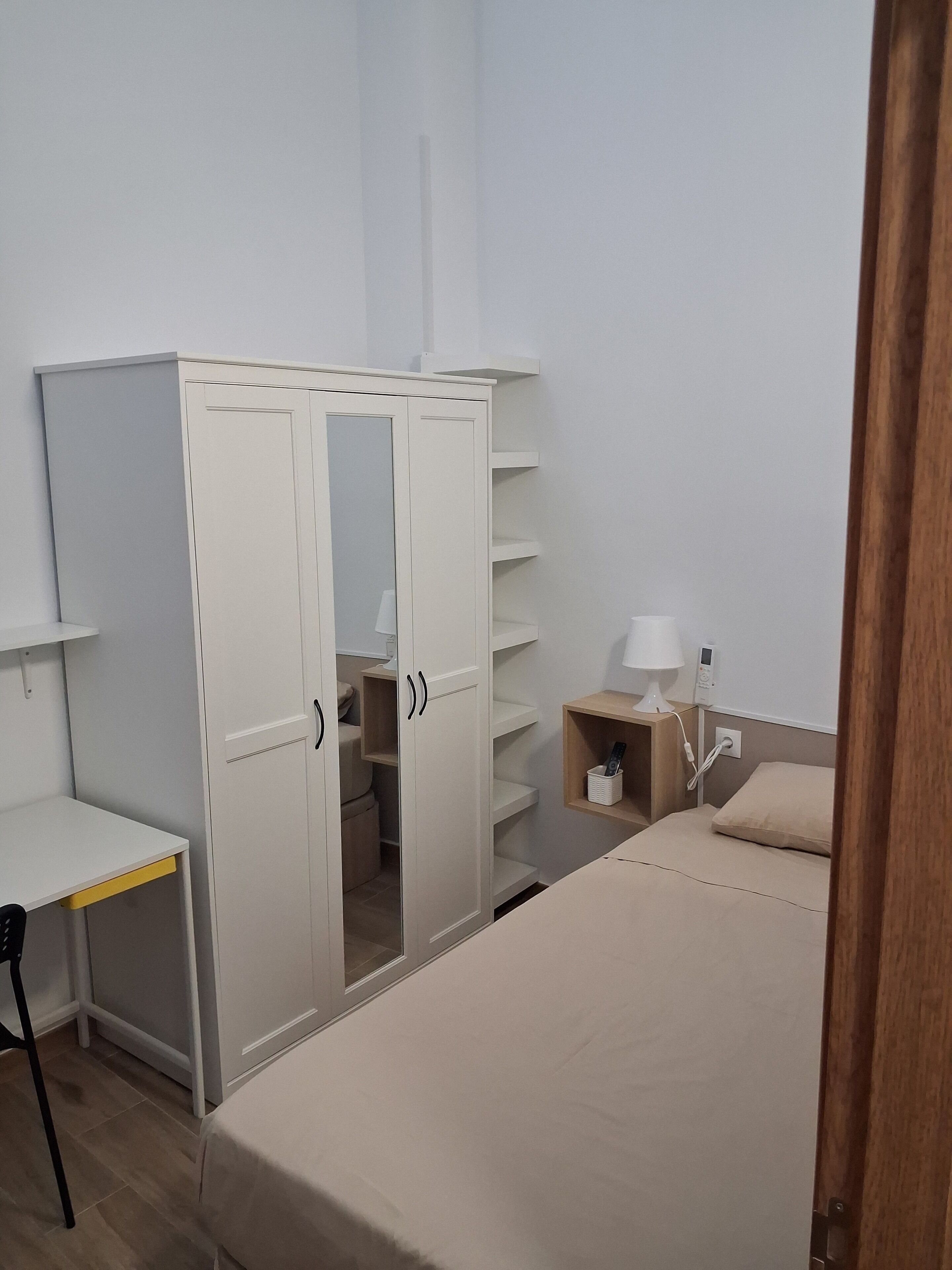 Een slaapkamer met een bed, een kledingkast met deuren, een nachtkastje met lamp en een klein plankje.