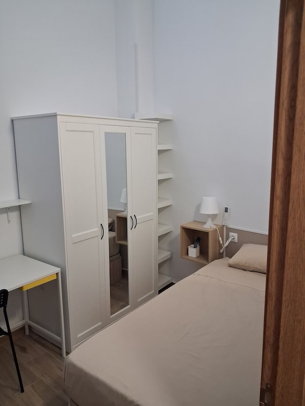 A bedroom with a bed, a wardrobe with multiple doors, a bedside table with a lamp, and a small shelf.