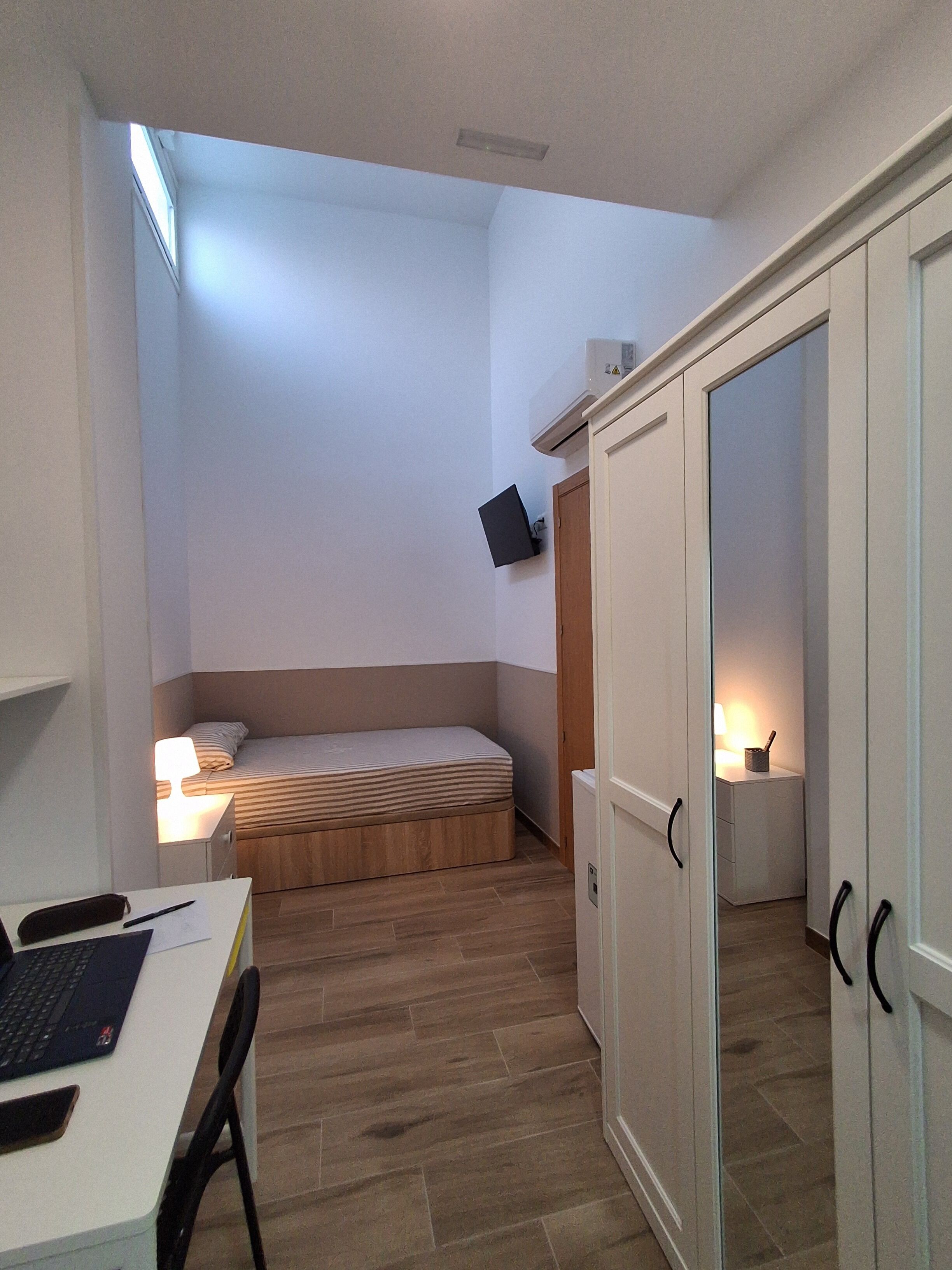 A compact room with a bed, a desk with a laptop, a wardrobe, and a bedside lamp.