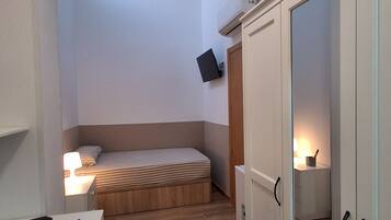 A compact room with a bed, a desk with a laptop, a wardrobe, and a bedside lamp.
