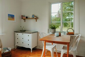 Ein Zimmer mit einem Holzesstisch, weißen Stühlen, einer weißen Kommode, einem Fenster mit Blick auf Bäume und einem Regal mit Büchern und Dekoration.