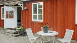 Ett trähus med en vit dörr, ett fönster och ett runt bord med två stolar. På bordet står det krukväxter och en lykta.
