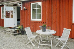 Ein Holzhaus mit weißer Tür, einem Fenster und einem runden Tisch mit zwei Stühlen. Auf dem Tisch stehen Blumentöpfe und ein Laternen.