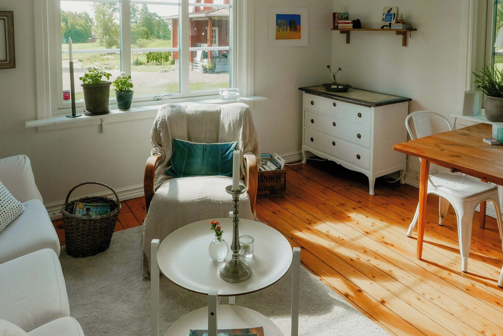 A cozy living room with a white sofa, a round table with a candle, a wooden dining table, and a white dresser with drawers.
