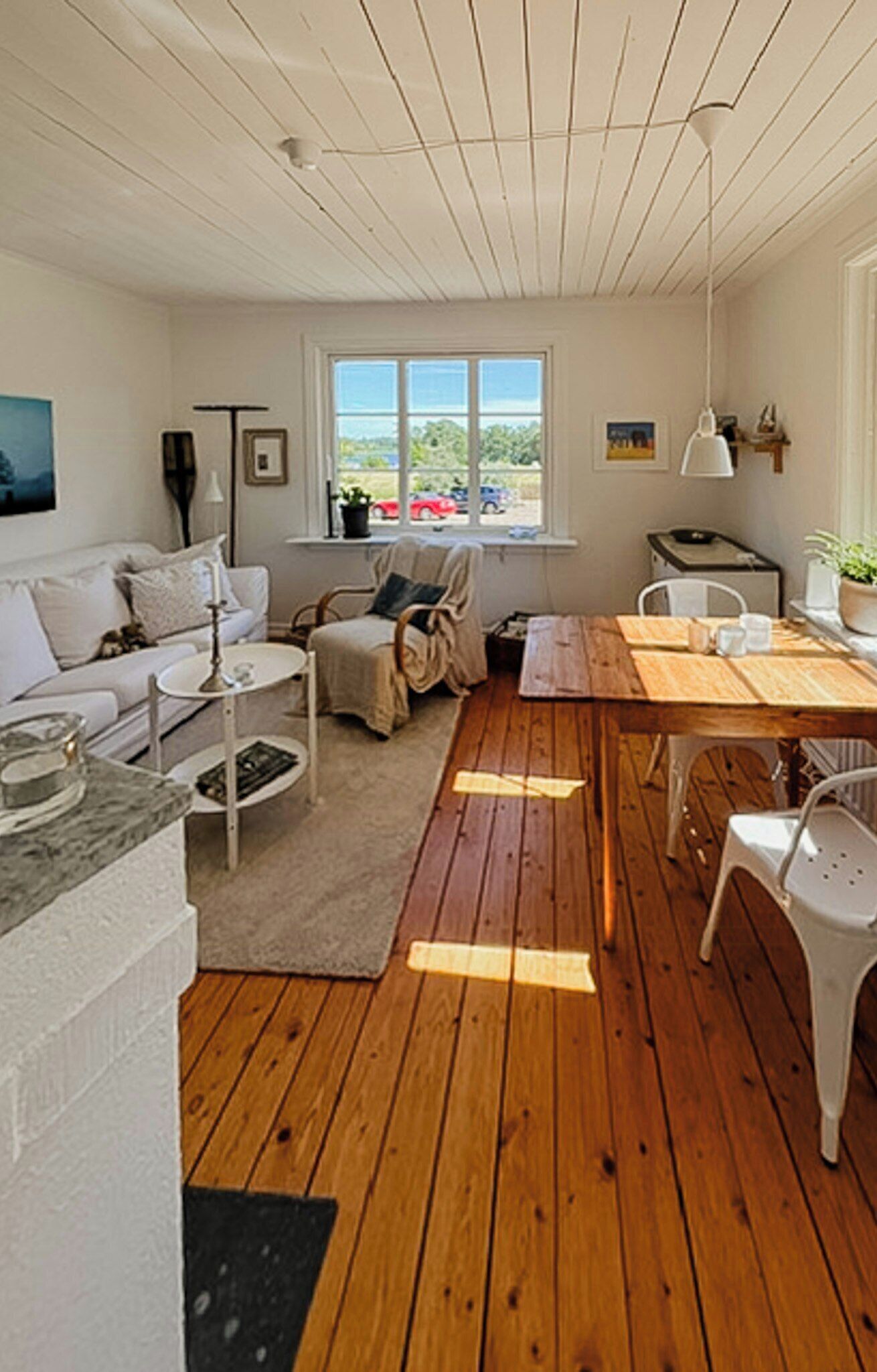 Un salón acogedor con suelo de madera, un sofá blanco, una mesa de comedor y una ventana con vistas a coches.