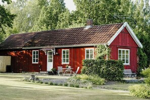 A red wooden house with a sloped roof, a front porch, and a small garden area with a bench and potted plants.