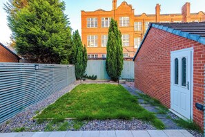 Un jardin à l’arrière d’une maison, avec une pelouse, une clôture en bois et un bâtiment en briques en arrière-plan.