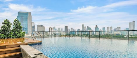 Una piscina infinita en la azotea con vistas a un horizonte urbano, una terraza de madera y una barandilla metálica.