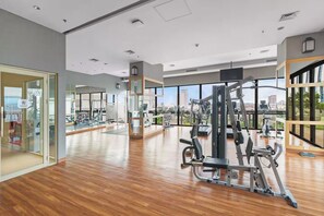 A modern gym with wooden floors, various exercise equipment, large windows offering city views, and a glass door entrance. - Brayen by Kozystay - Tebet (South Jakarta)