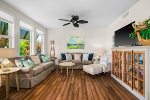 A living room with a sectional sofa, a round coffee table, a television on a cabinet, and a ceiling fan. - Pili Mai 6F by Alekona Kauai - 2BR Condo access to fitness (Koloa)