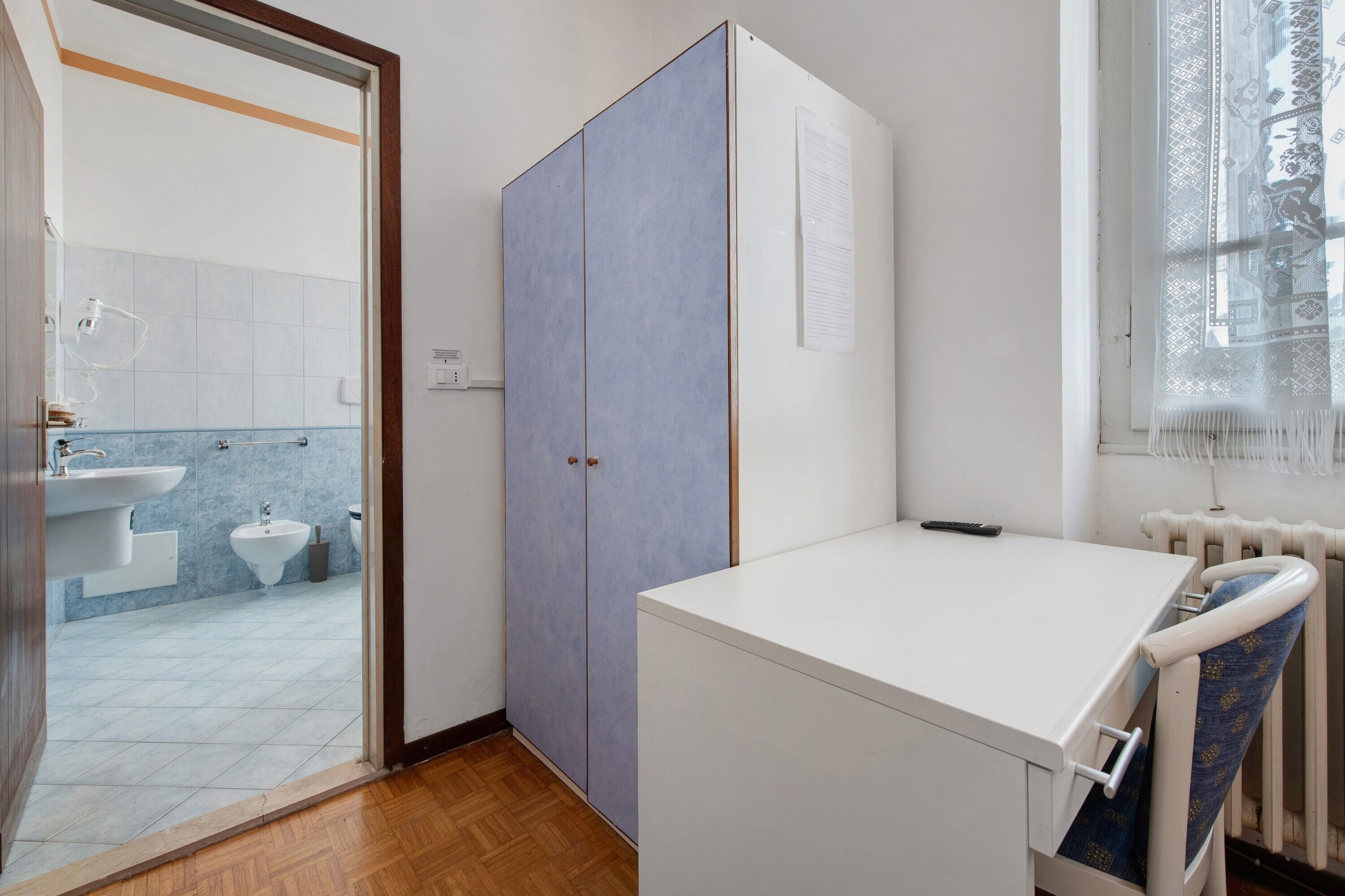 A small, compact room with a white desk, a radiator, and a window with a curtain.