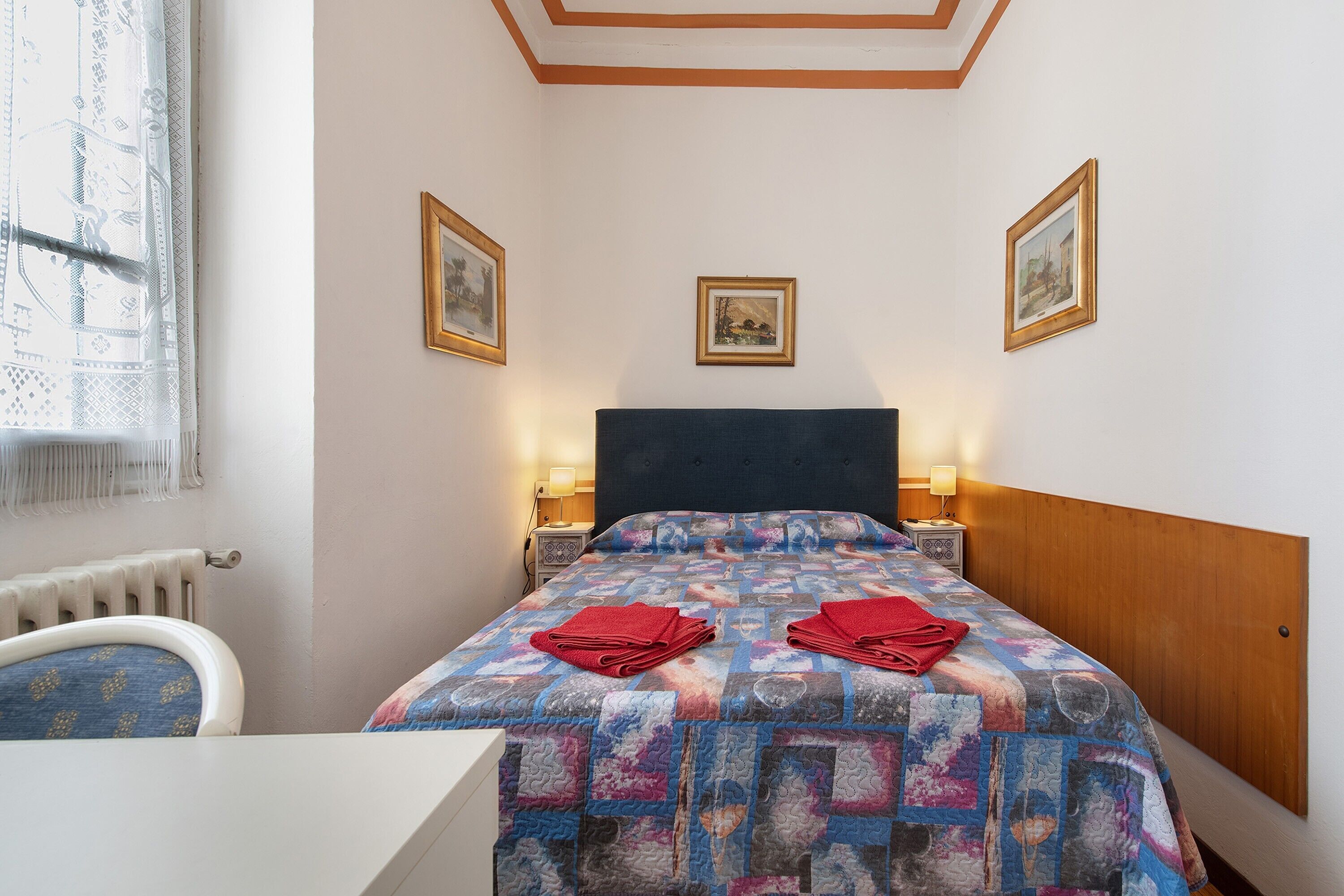 A hotel room with a bed, two bedside lamps, a window with curtains, and three framed pictures on the wall.