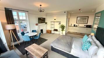 A modern hotel room with a bed, a sofa, a dining table, and a kitchen area.