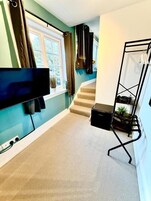 Una habitación con un televisor de pantalla plana montado en la pared, una ventana con cortinas, una escalera y un soporte de metal negro con objetos encima.