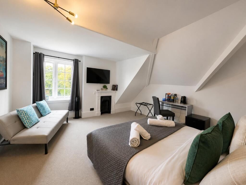 A modern hotel room with a large bed, a sofa, a desk with a chair, a flat-screen TV, and a fireplace.