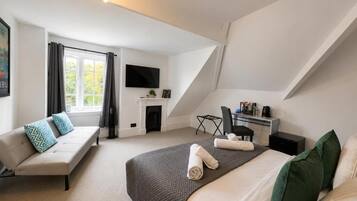 A modern hotel room with a large bed, a sofa, a desk with a chair, a flat-screen TV, and a fireplace.