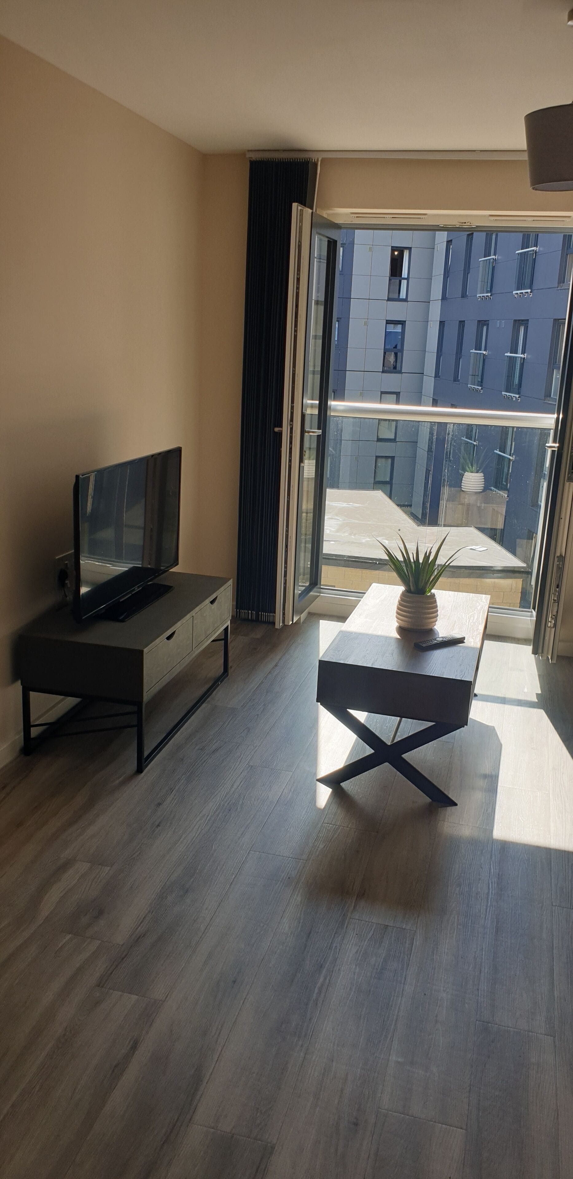 En moderne stue med et fladskærms-tv, et sofabord og udsigt til en bygning gennem en glasdør.