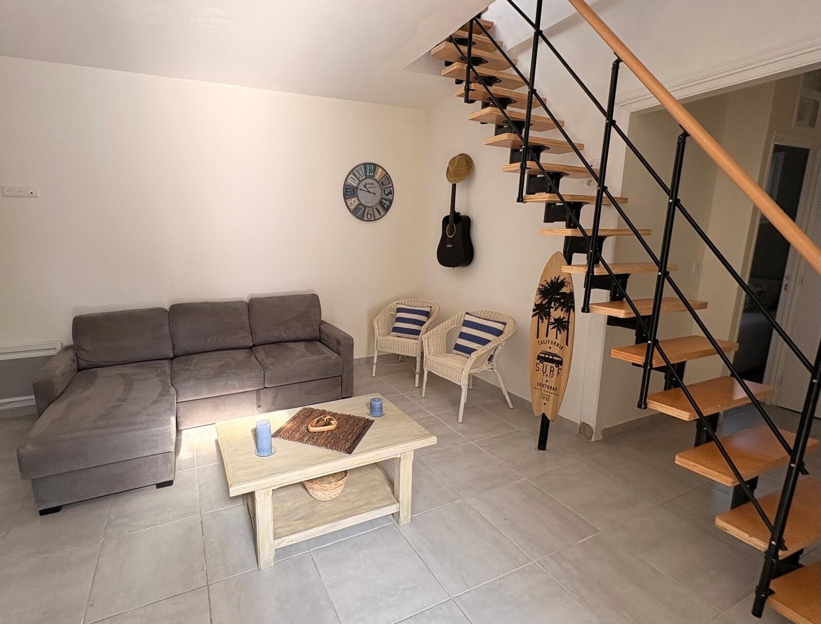 A living room with a grey sofa, two wicker chairs, a coffee table, a surfboard decoration, a clock, and a spiral staircase.