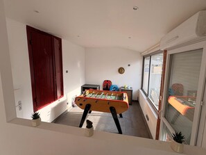 Ein schmales Zimmer mit einem Kicker, einer roten Tür und einem Fenster mit Blick auf die Straße.