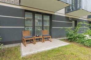 Eine Terrasse mit zwei Holzstühlen, einem kleinen Tisch, einer Glastür und einem grünen Busch.