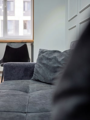 A grey sofa with a cushion, a black chair, and a window with a view of buildings.