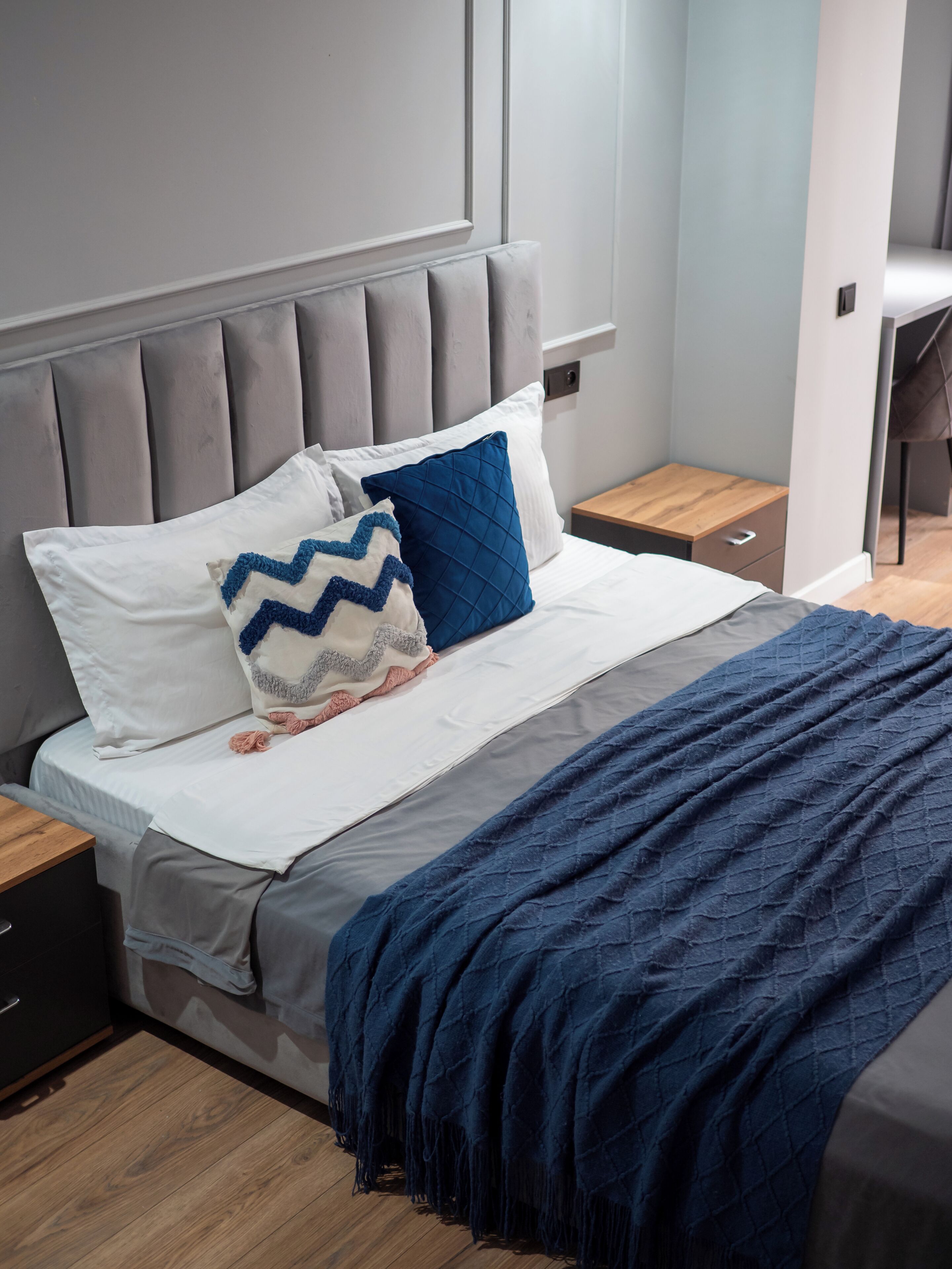 Ein modernes Schlafzimmer mit grauem gepolsterten Kopfteil, einem Bett mit weißblauer Bettwäsche und einer dunkelblauen Tagesdecke.