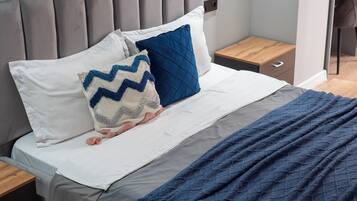 Ein modernes Schlafzimmer mit grauem gepolsterten Kopfteil, einem Bett mit weißblauer Bettwäsche und einer dunkelblauen Tagesdecke.