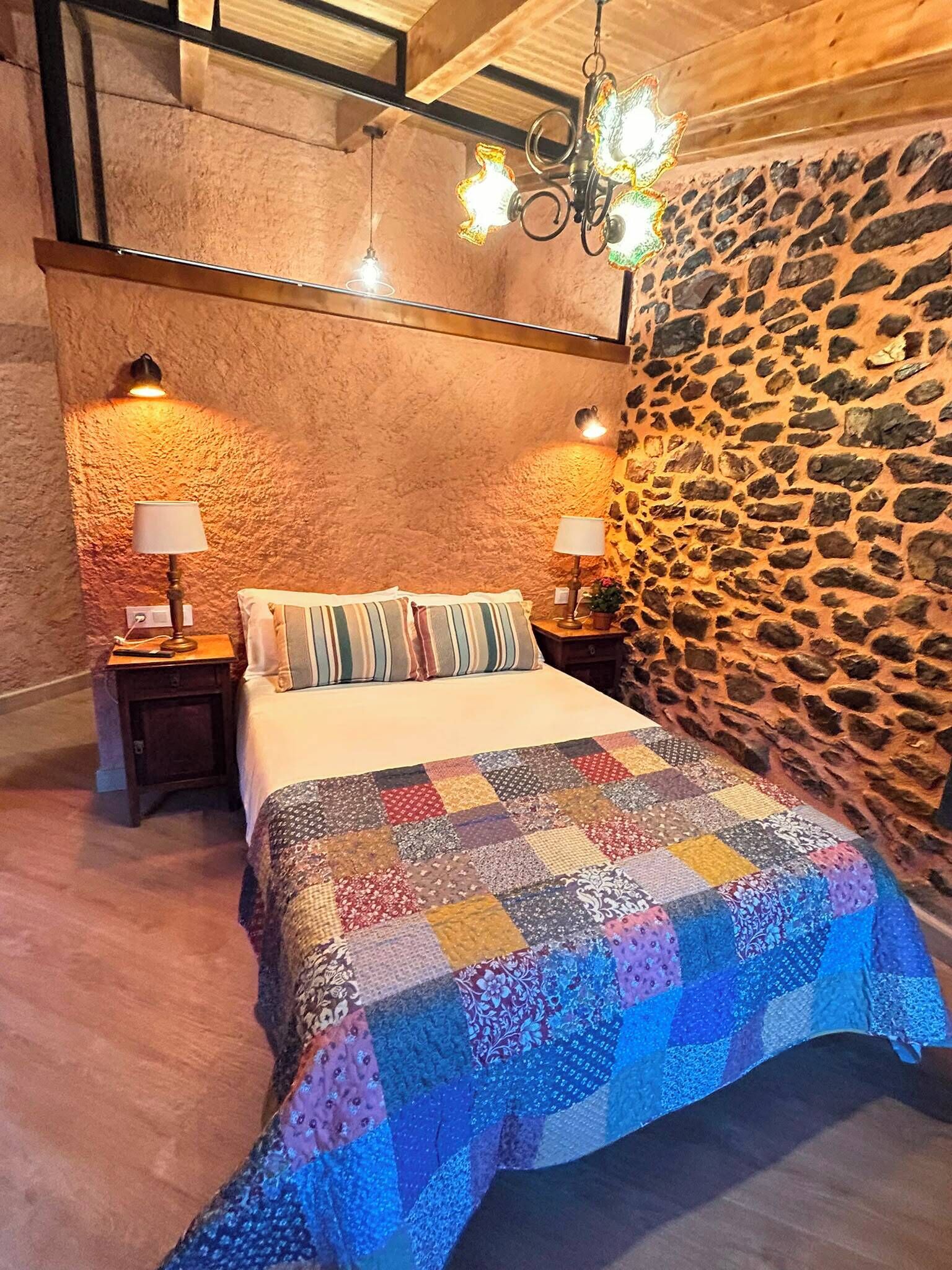 A bedroom with a stone wall, a quilted bedspread, and two bedside lamps.