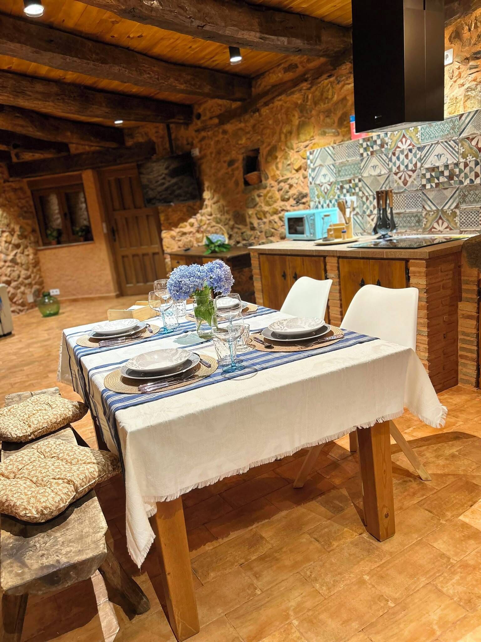 A dining table set with plates, glasses, and a vase of flowers, surrounded by stone walls and a tiled backsplash.
