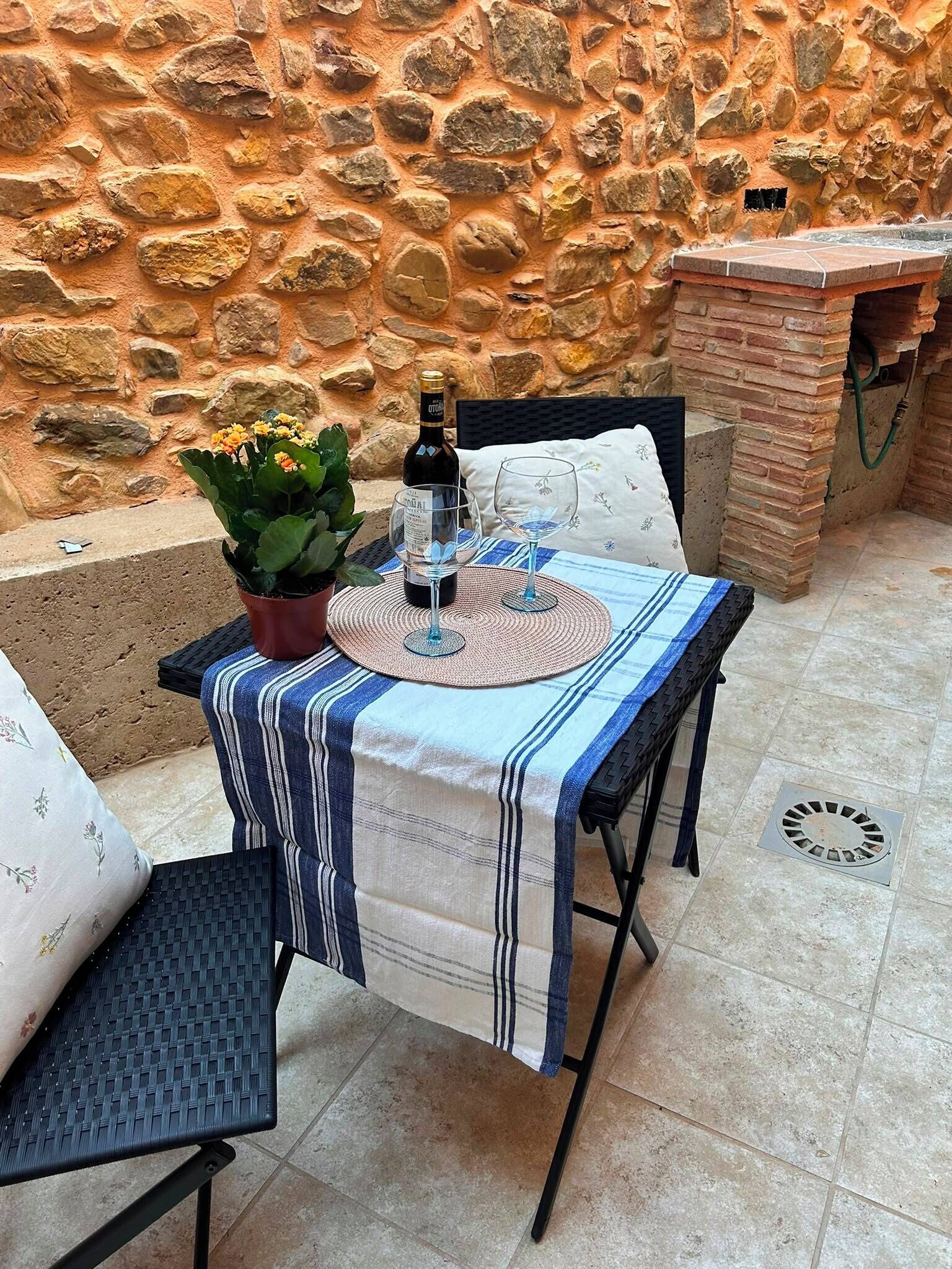 Een terras buiten met een tafel gedekt voor twee personen, een potplant en een fles wijn.