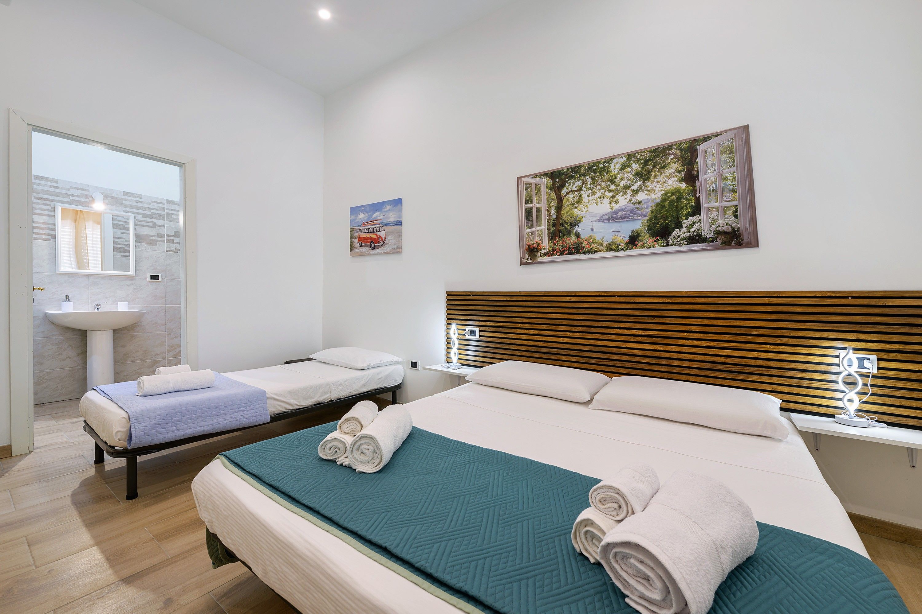 A hotel room with two single beds, a large window with a view, a painting on the wall, and a bathroom with a sink and mirror.