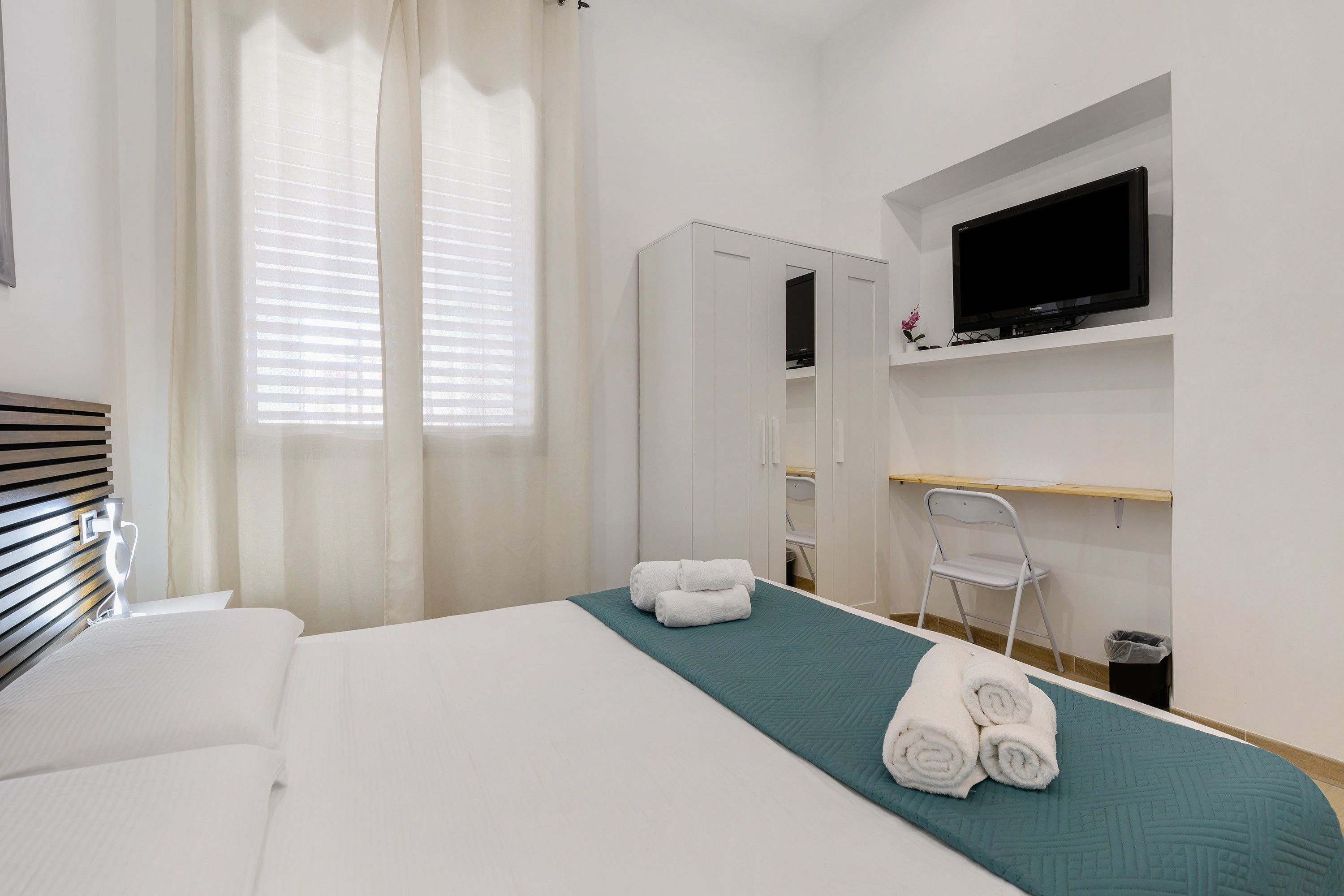 Ein Hotelzimmer mit einem Bett, einem an der Wand befestigten Fernseher, einem Spiegel und einem kleinen Schreibtisch mit Stuhl.
