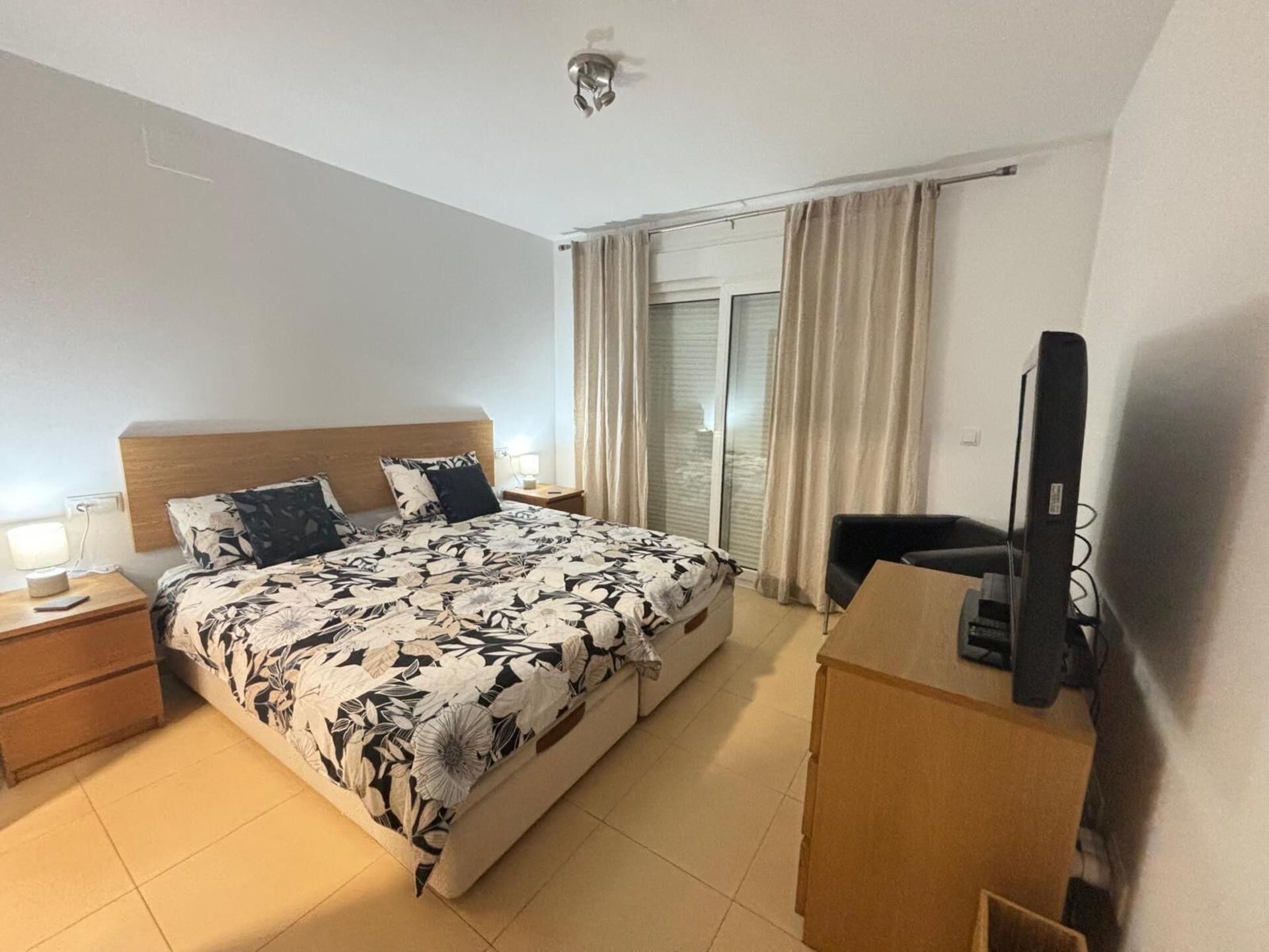A hotel room with a bed, bedside tables, a TV, and a window with curtains.